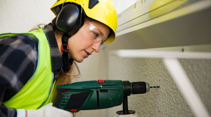 Young Female construction worker in hard hat drilling concrete wall with a drill and smiling at the camera. Building and renovation. feminism concept